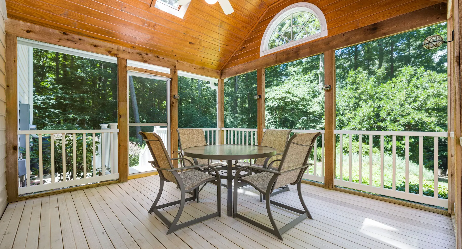 Screened-in covered porch with vaulted wood ceiling, dining table, and panoramic forest views through Mirage retractable screens